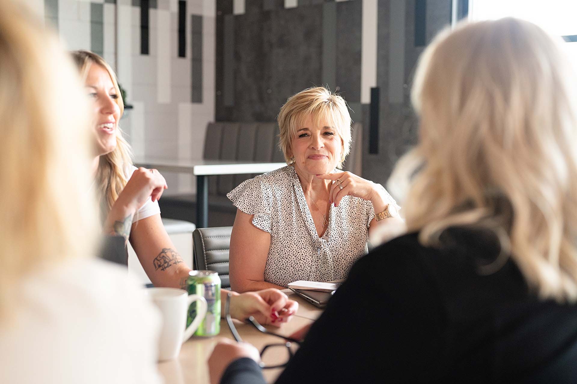 Group of professional women having a meeting around a table, with a focus on a woman in a light patterned blouse, thoughtfully engaged in the conversation.