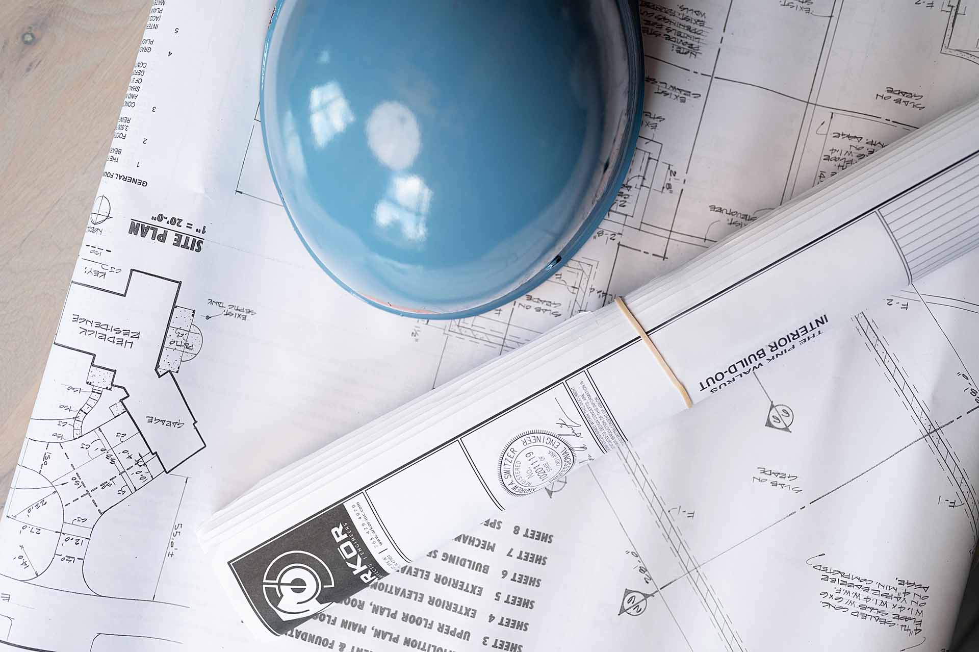 A close-up of construction blueprints laid out on a table, featuring detailed site and building plans. A blue hard hat is placed on top of the blueprints, symbolizing construction and planning. A rolled set of architectural drawings secured with a rubber band is visible, emphasizing the meticulous nature of the construction and design process.