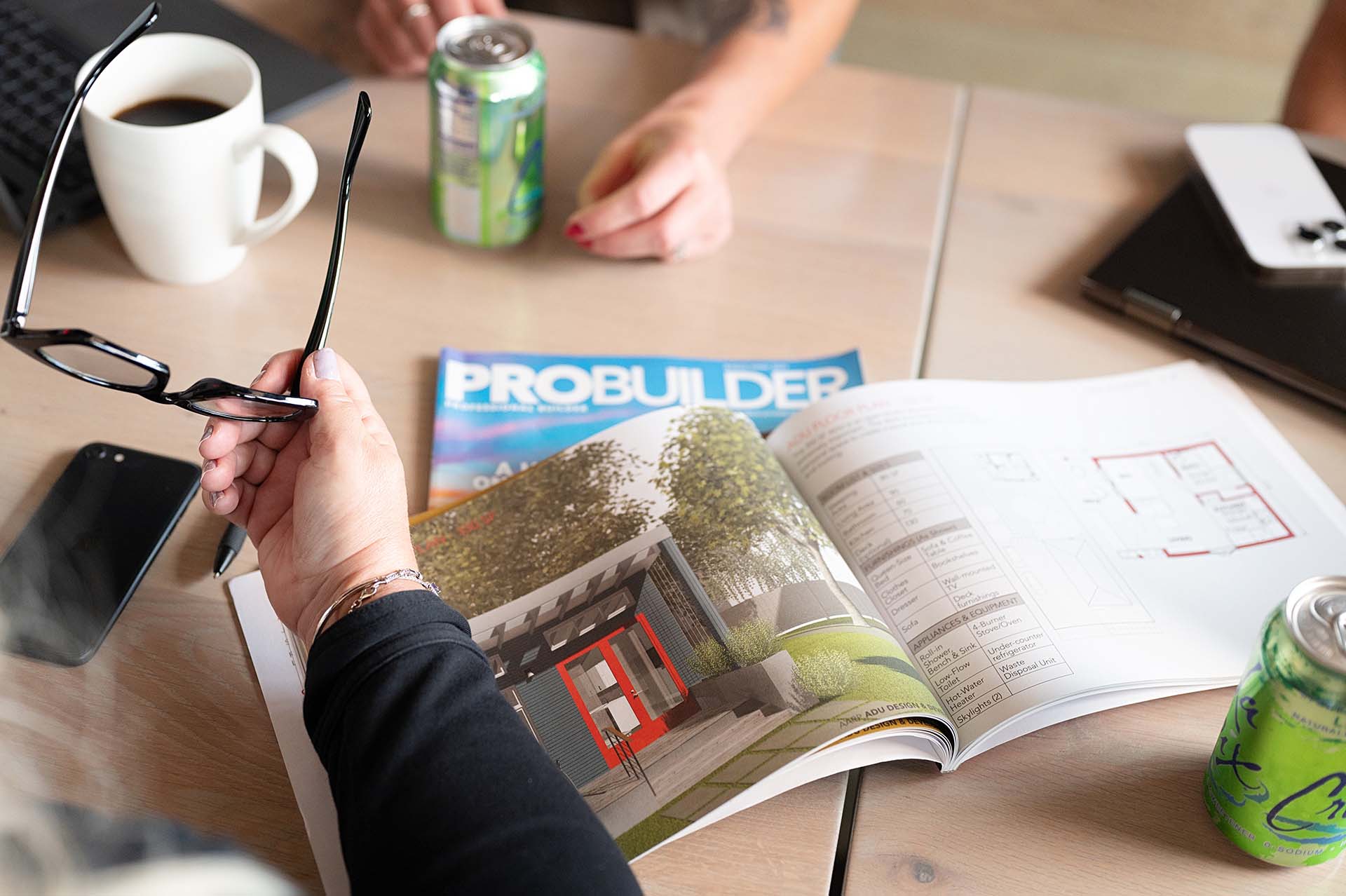 A desk with an open 'Probuilder' magazine showing a modern house design. Visible are hands holding glasses, a white coffee mug, green beverage cans, a smartphone, and papers. The scene suggests a casual work or planning session focused on architecture or construction.